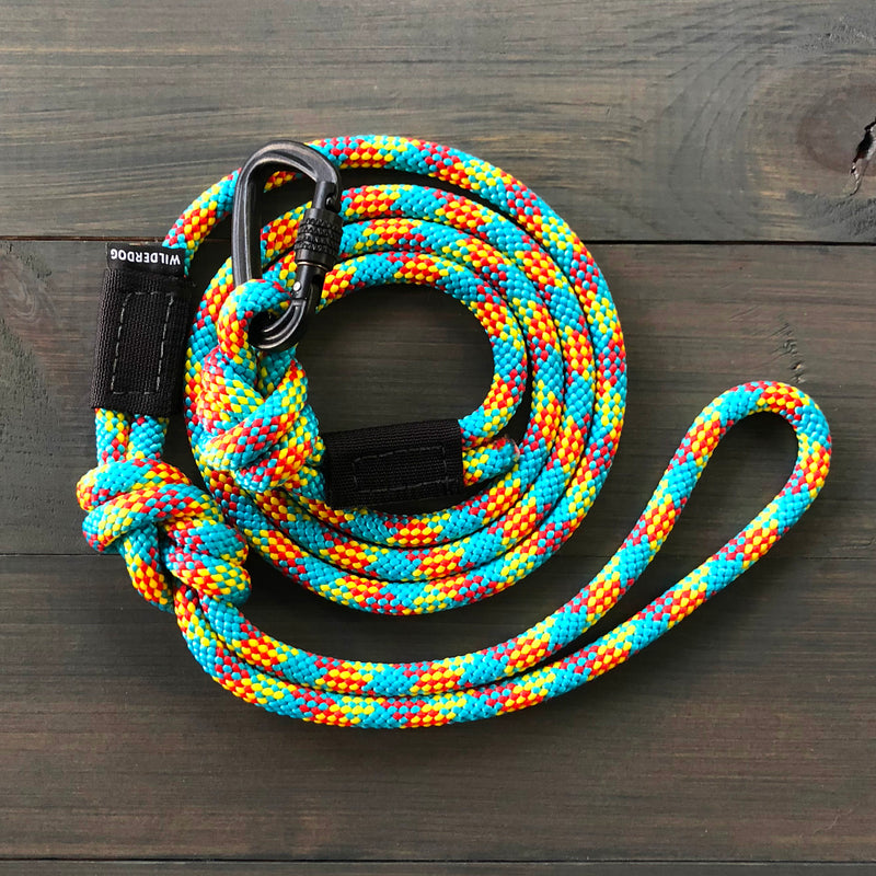 A vibrant, multi-colored Aprés Small Carabiner Leash crafted from rock climbing rope features blue, orange, yellow, and white patterns coiled neatly on a dark wooden surface. The leash includes a black metal clip and a black label with white text reading "Wildebeest.